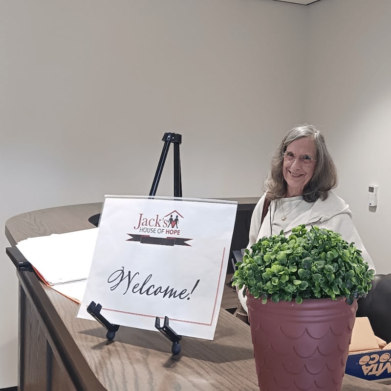 A smiling woman stands behind a wooden counter featuring a "Welcome!" sign and a potted green plant against a plain white wall.