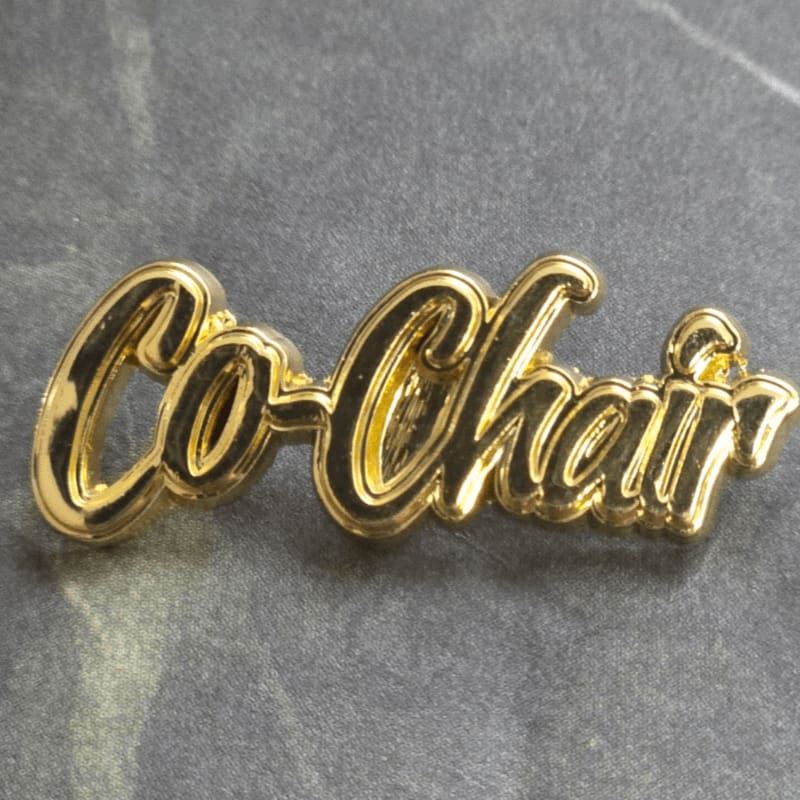 A gold-colored "Co-Chair" pin rests on a textured grey surface.