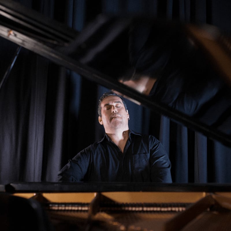 A man with closed eyes plays a piano, framed by the instrument's lid in the foreground and dark curtains in the background.