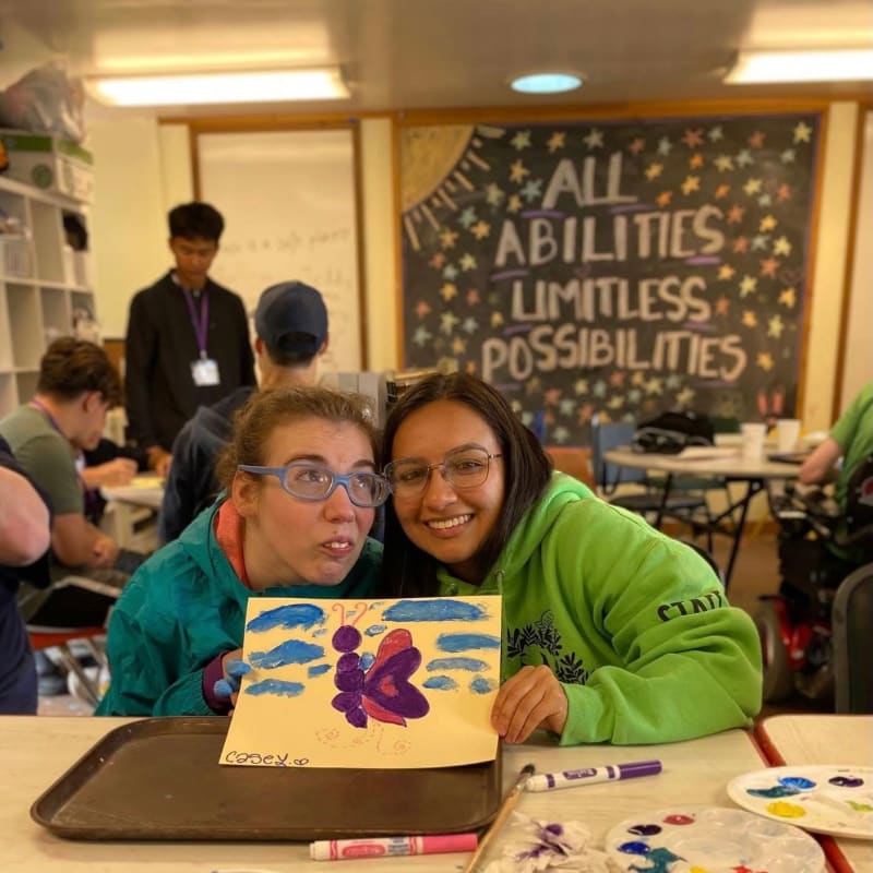 Two smiling women pose with a colorful painting in the foreground, while other students and an inspirational chalkboard are visible in the background classroom.
