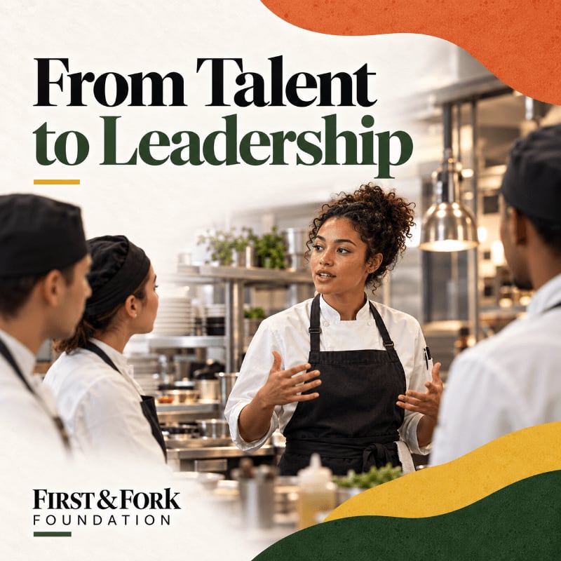 A female chef in a white coat and black apron speaks to her colleagues in a professional kitchen setting.