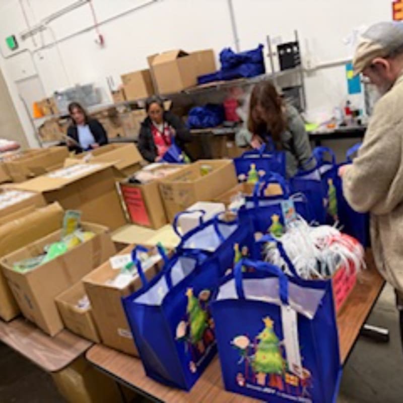 Blue gift bags and cardboard boxes fill the foreground, while several people organize items in a warehouse setting in the background.