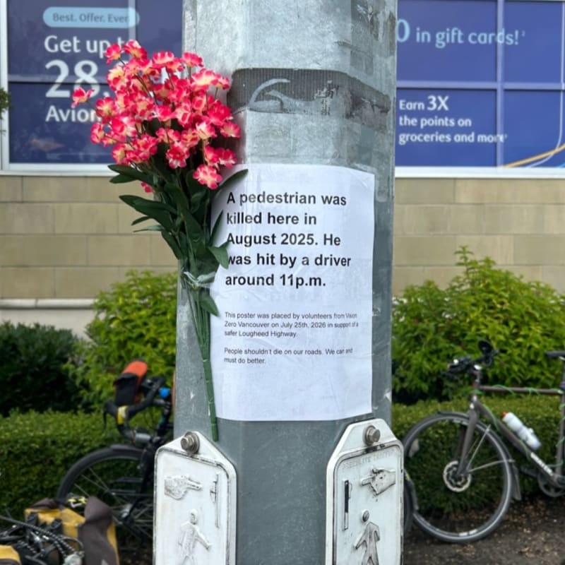 A memorial with flowers and a notice about a pedestrian fatality is attached to a pole in front of a store with bicycles parked nearby.