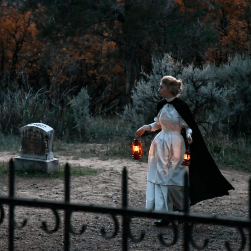A woman in a white dress and black cloak holds lanterns in a graveyard, with a headstone in the foreground and a dark, wooded forest in the background.