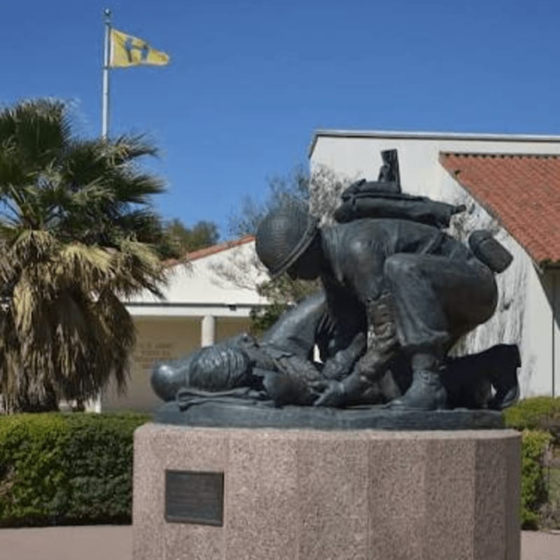 A bronze sculpture of a soldier tending to a fallen comrade sits on a stone pedestal, with a palm tree, a white building, and a yellow flag in the background.