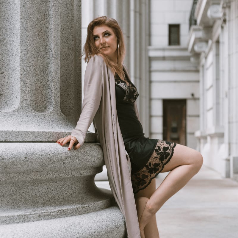 A woman in a black lace dress and long beige cardigan leans against a large stone column in front of a classical building.