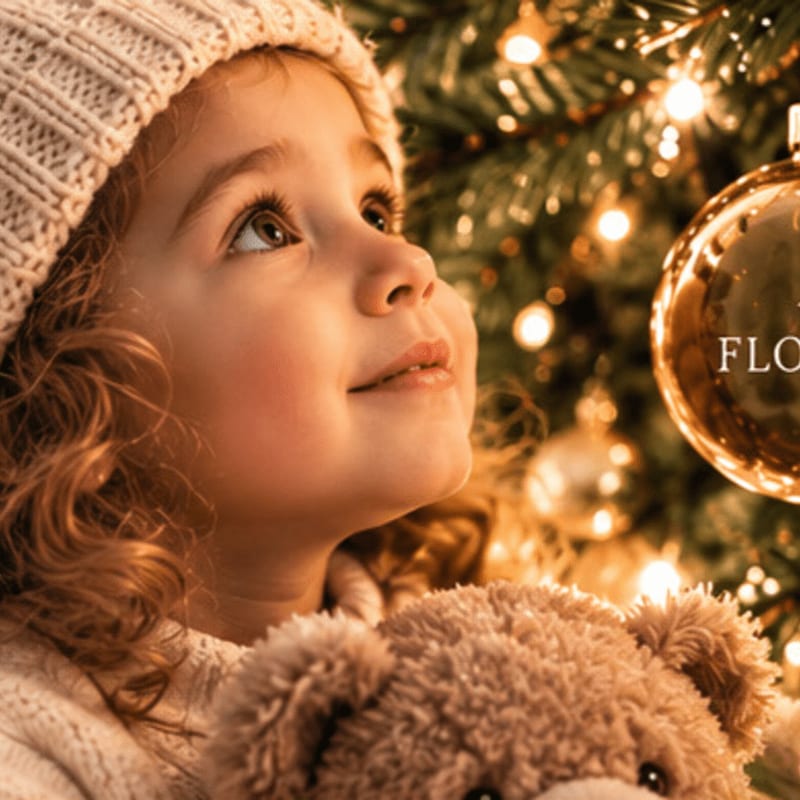 A young girl in a knit hat holds a teddy bear in the foreground, gazing up at a glowing, decorated Christmas tree in the background.