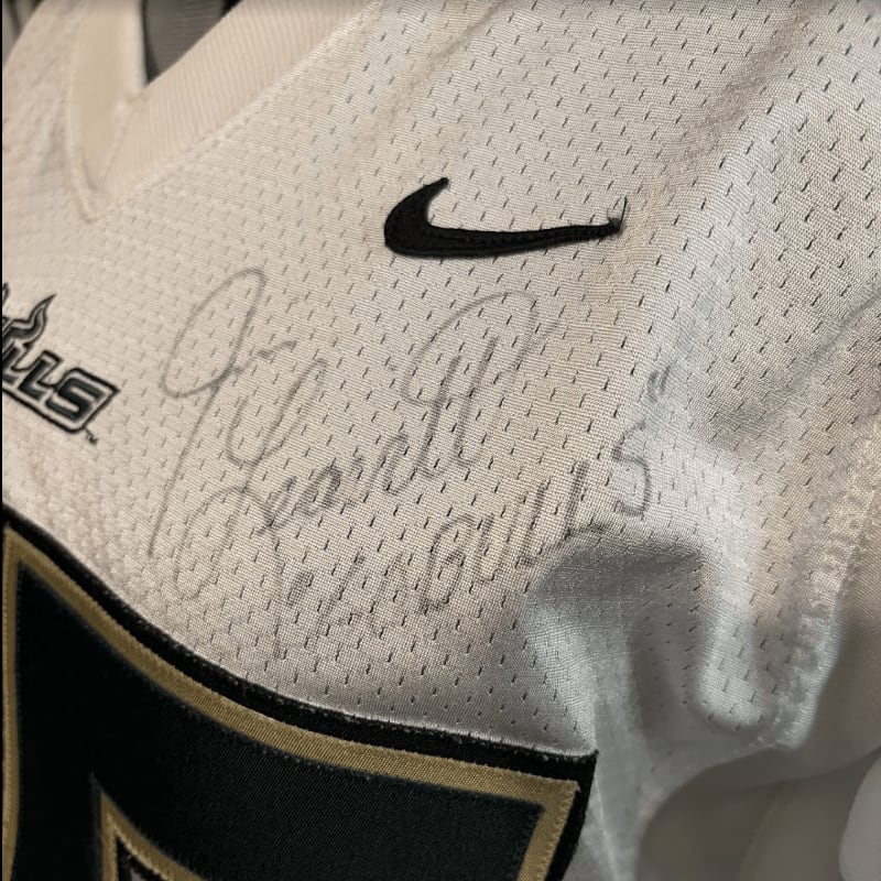 A black Nike swoosh and a handwritten signature are featured on the foreground of a white mesh jersey against a blurred white fabric background.