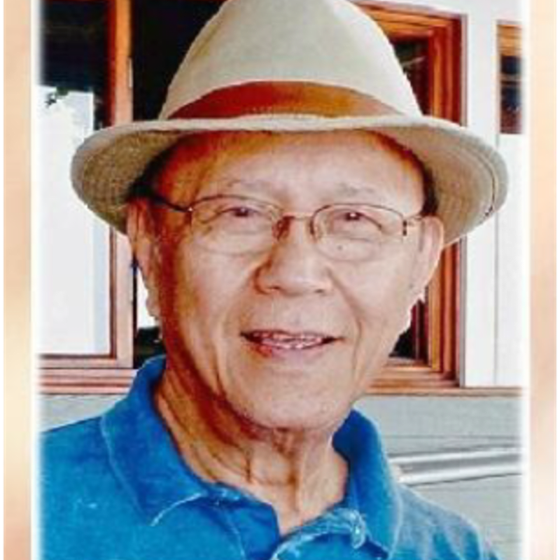 An elderly man wearing glasses, a tan hat, and a blue polo shirt stands in front of a building with wooden window frames.