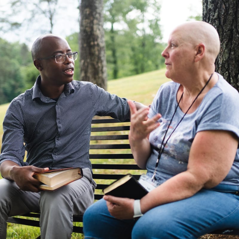A man and a woman hold books while talking on a park bench, with green grass and trees in the background.