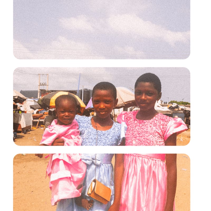 Three young girls in pink and blue dresses stand together in the foreground against a blurred outdoor market background.