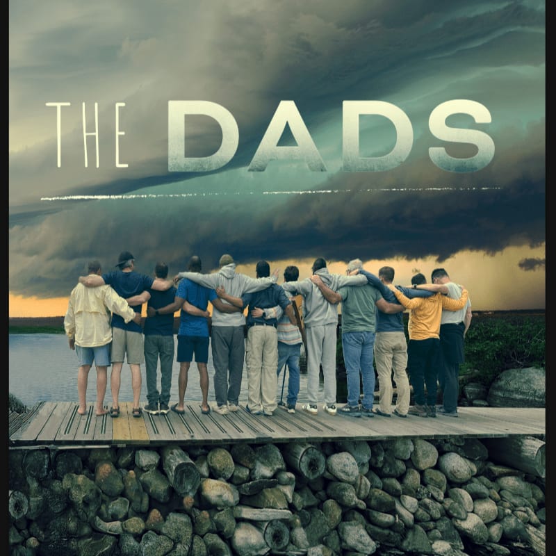 A group of men stand with their arms around each other on a wooden pier, set against a backdrop of a dark, stormy sky and a calm body of water.