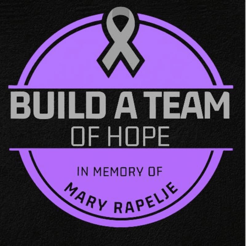 A purple circular emblem featuring a grey ribbon and the text "BUILD A TEAM OF HOPE IN MEMORY OF MARY RAPELJE" is set against a black textured background.