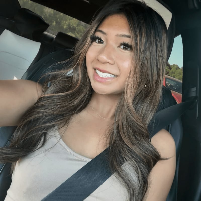 A smiling woman with long, wavy brown hair sits in the driver's seat of a car, with a blurred outdoor landscape visible through the windows.