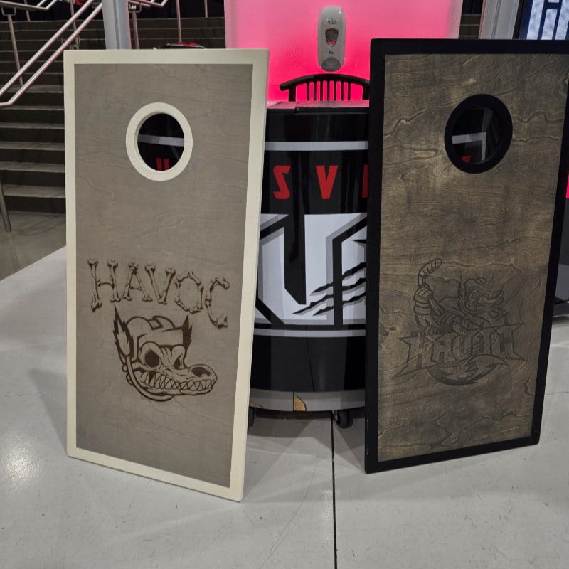 Two cornhole boards with "Havoc" branding stand in the foreground against a background featuring a black barrel and a staircase.