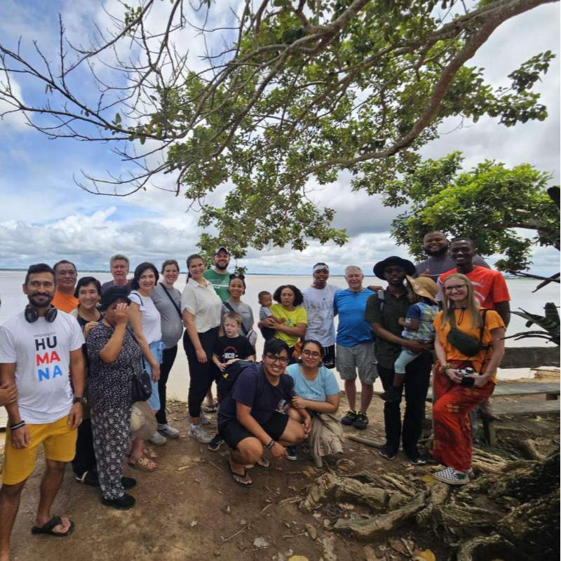 A diverse group of people poses for a photo under a large leafy tree with a cloudy sky and a body of water in the background.