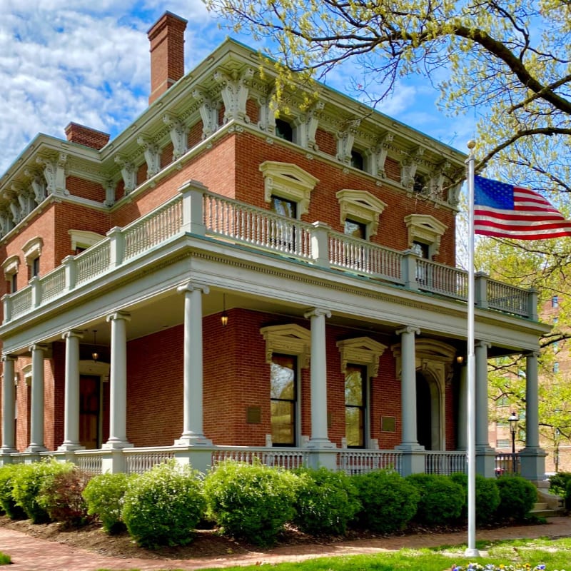 A red brick mansion with a white porch and an American flag stands before a bright blue sky and leafy trees.