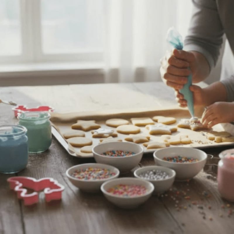 A person decorates star-shaped cookies with blue icing on a wooden table surrounded by bowls of colorful sprinkles and cookie cutters, with a bright window in the background.