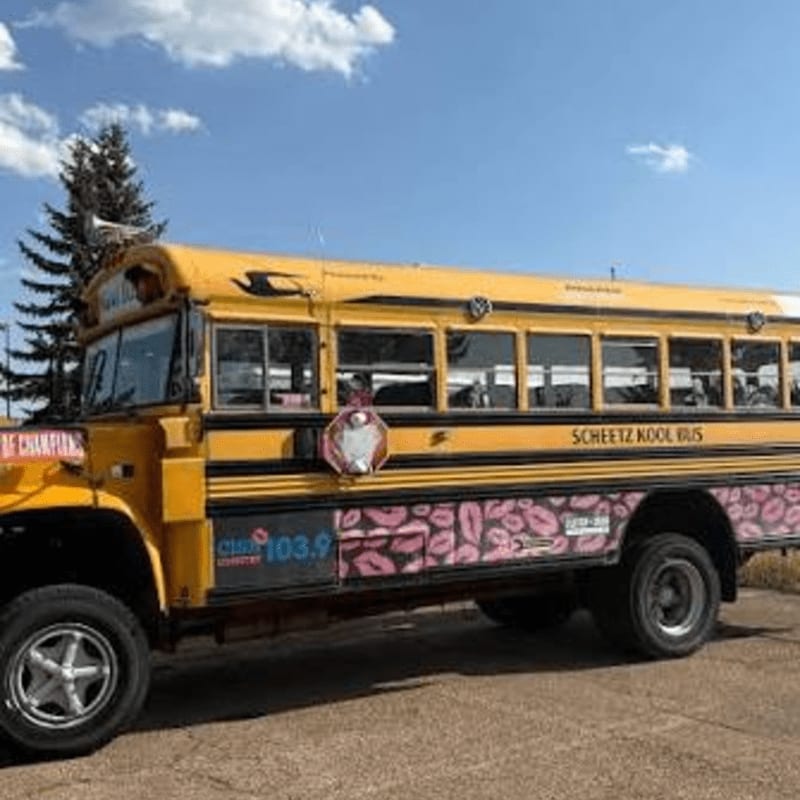 A yellow school bus with pink lip decals sits on a paved surface against a backdrop of a green tree and a blue sky with white clouds.