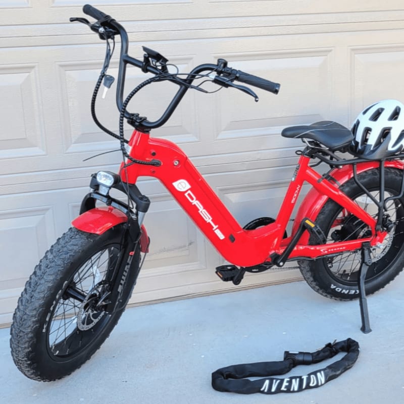 A red electric bike and a black strap sit on a concrete driveway in front of a white garage door.