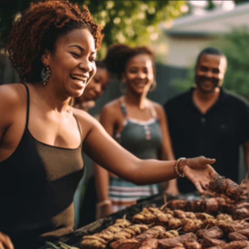 A smiling woman grills meat in the foreground while friends watch and smile in the blurred outdoor background.