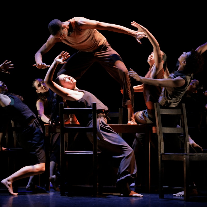 A group of dancers in dark clothing perform an expressive routine with chairs against a stark black background.