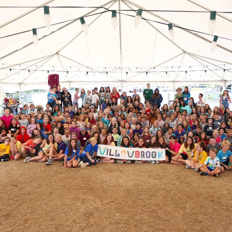 A large group of children and adults pose for a photo under a white tent, with a "WILLOWBROOK" banner in the foreground and a grassy field in the background.