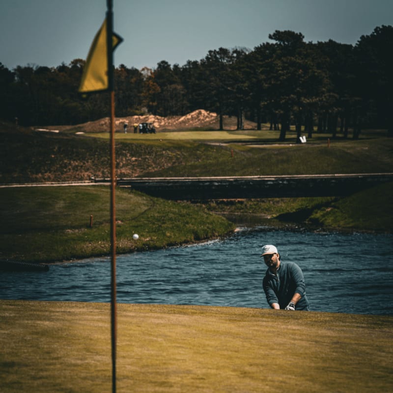 A yellow flag stands in the foreground of a golf course, with a man standing by a water hazard and a backdrop of rolling hills and dense trees.