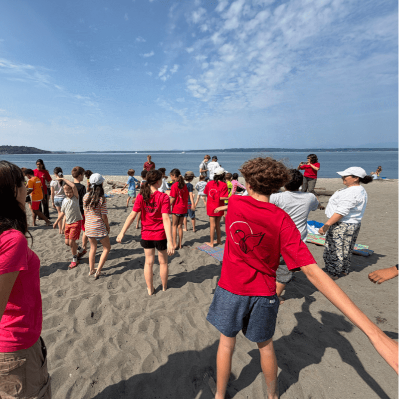 A group of children in red shirts gather on a sandy beach under a blue sky with a calm ocean in the background.