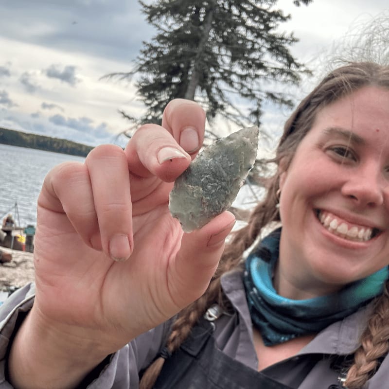 A smiling woman holds up a small, jagged stone in the foreground against a background of a lake, a pine tree, and a cloudy sky.