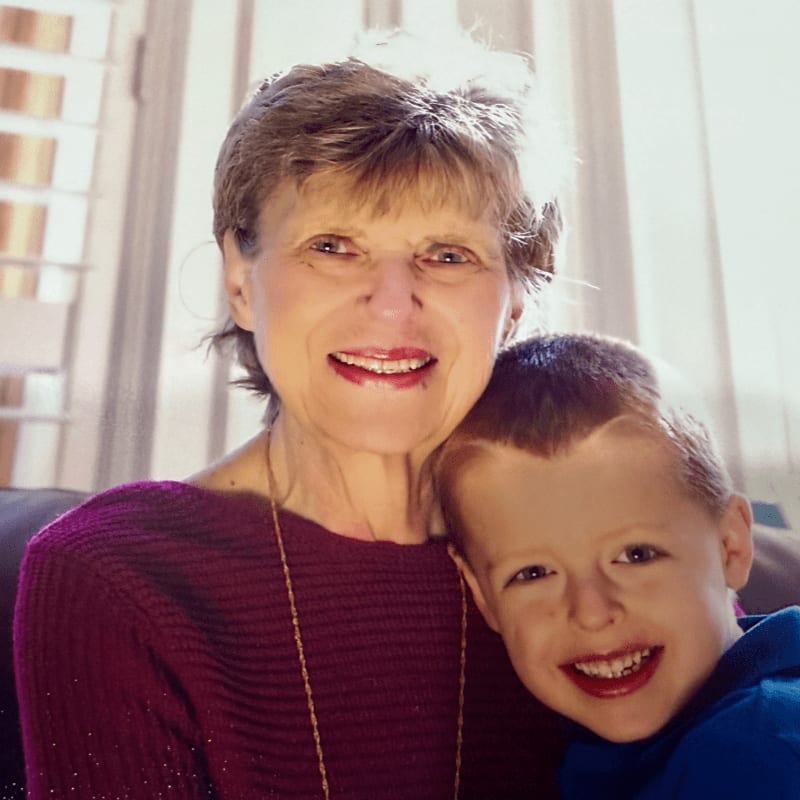 A smiling woman in a maroon sweater and a young boy in a blue shirt pose together against a bright, blurred indoor background.