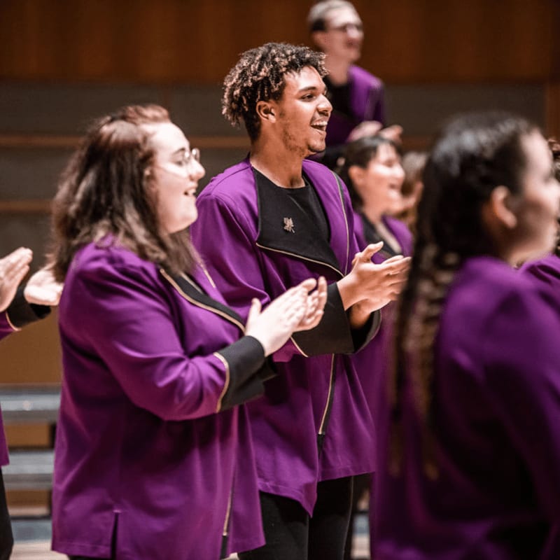 A group of people in purple robes are clapping and smiling in the foreground against a blurred wooden background.