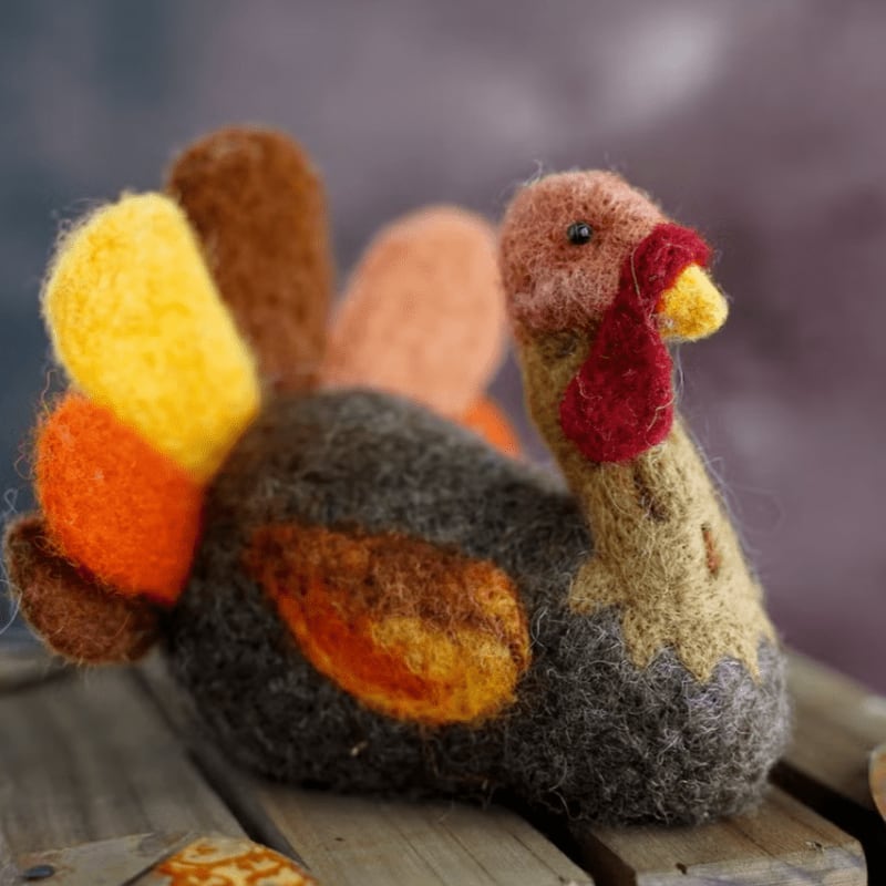 A colorful felted turkey sits on a wooden surface against a blurred purple background.