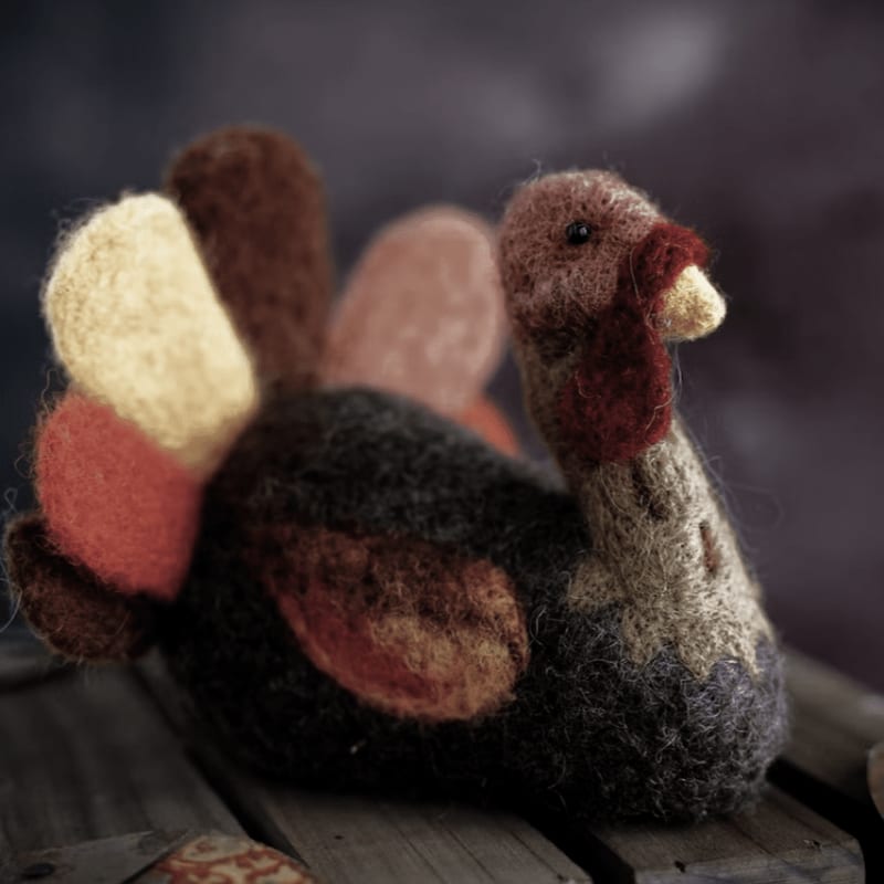 A colorful felted turkey sits on a wooden surface against a blurred purple background.