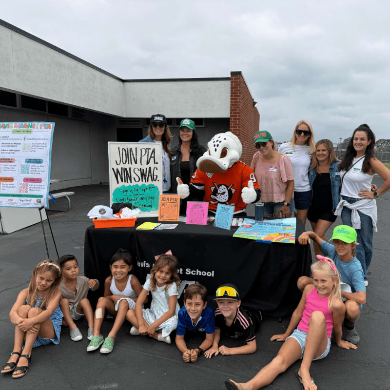 A group of adults and children pose behind and in front of a PTA promotional table in a paved outdoor area with a white building in the background.