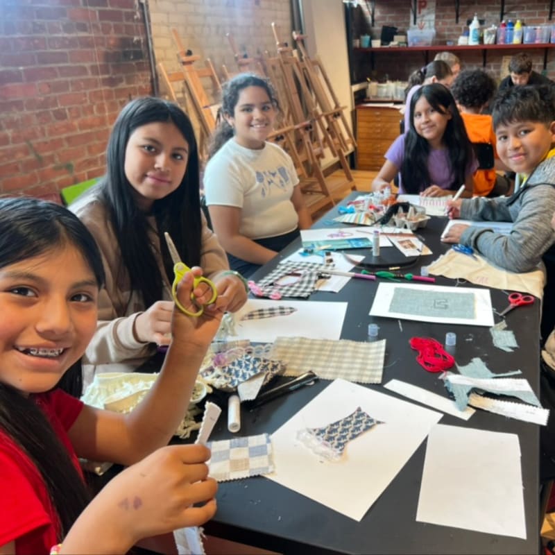 Children sit at a table covered in fabric scraps and craft supplies, with art easels and a brick wall in the background.