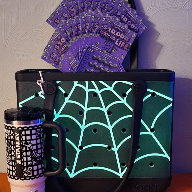 A glowing green spiderweb-patterned bag and a Stanley tumbler sit in the foreground against a background of purple lottery tickets and a textured wall.