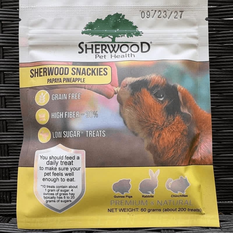 A bag of Sherwood Snackies Papaya Pineapple treats for small pets is shown against a dark, woven background, with a guinea pig eating a treat from a hand in the foreground.
