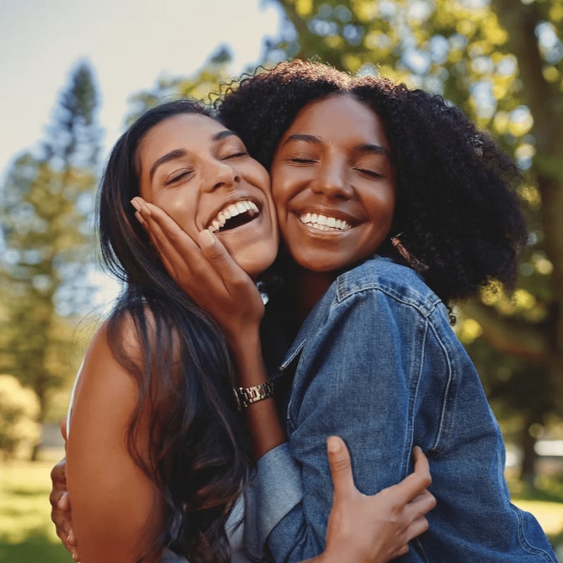 Two laughing women embrace in the foreground against a blurred background of green trees and sunlight.