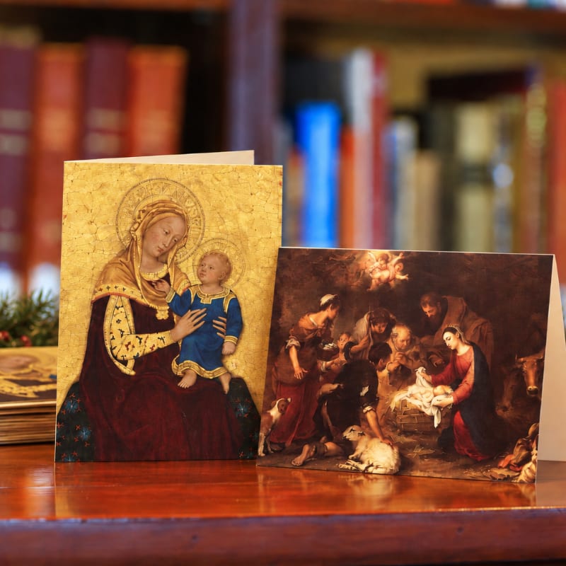 Two religious art cards sit on a wooden table in front of a blurred bookshelf.