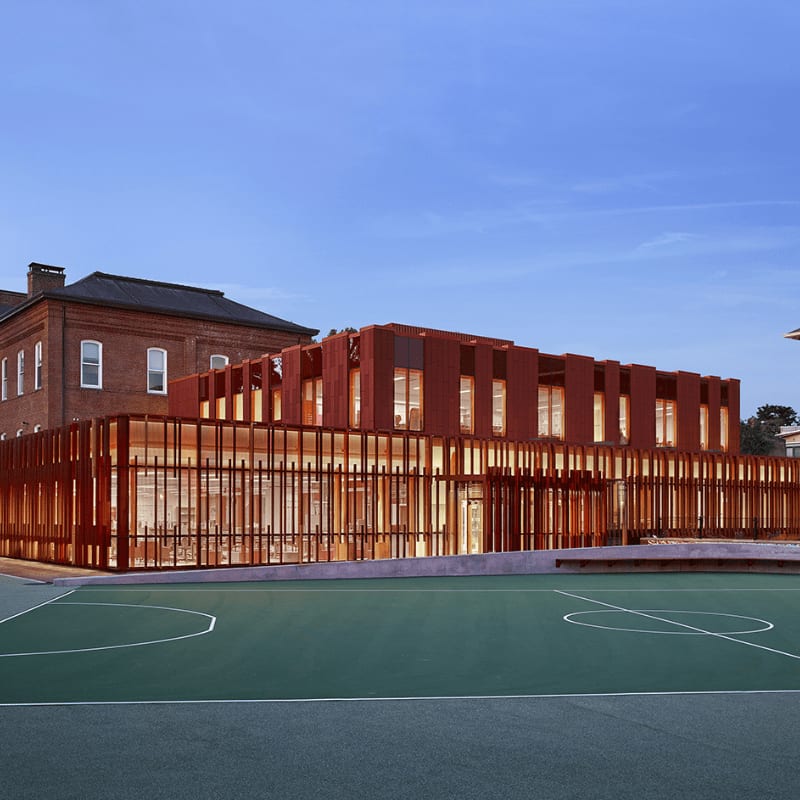 A green sports court sits in the foreground before a modern, rust-colored building with vertical slats, set against a clear blue sky and an older brick structure.