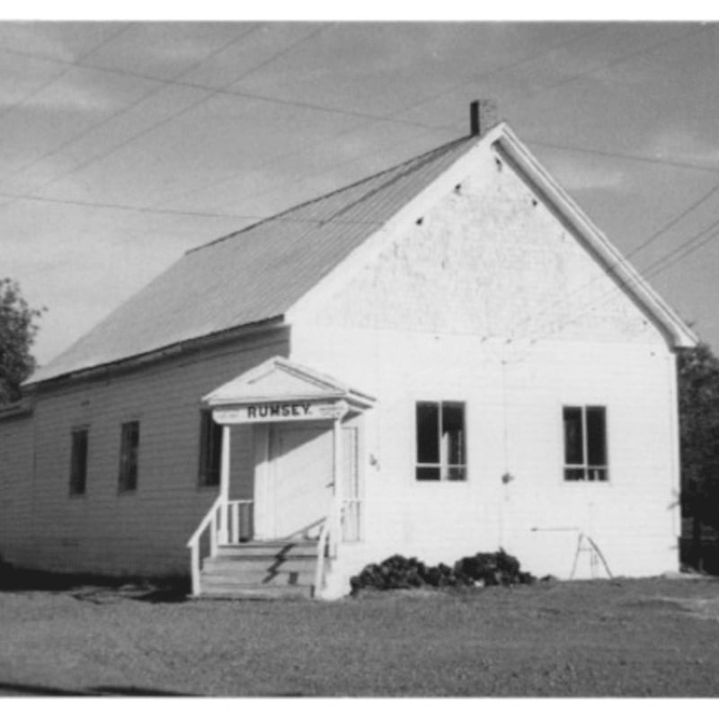 A white wooden building with a sign reading "RUMSEY" stands in the foreground against a clear sky with power lines in the background.