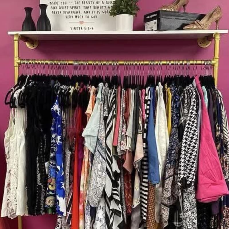 A variety of colorful patterned clothes hang on a gold rack beneath a white shelf holding vases, a plant, and shoes against a purple wall.