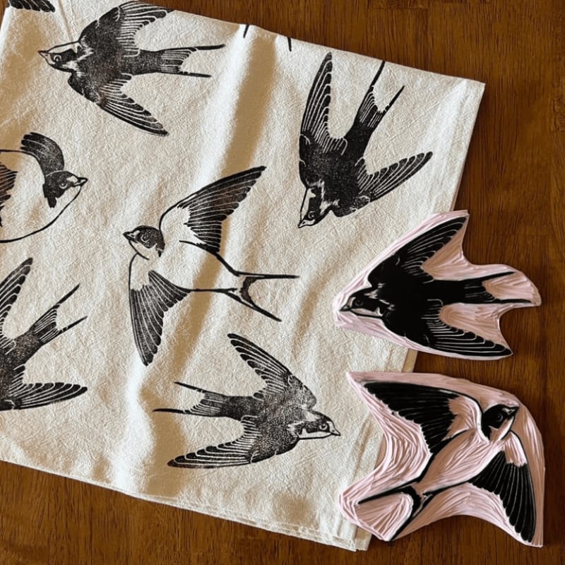 Two carved linoleum blocks of swallows sit atop a white fabric printed with black swallow designs, all resting on a wooden surface.