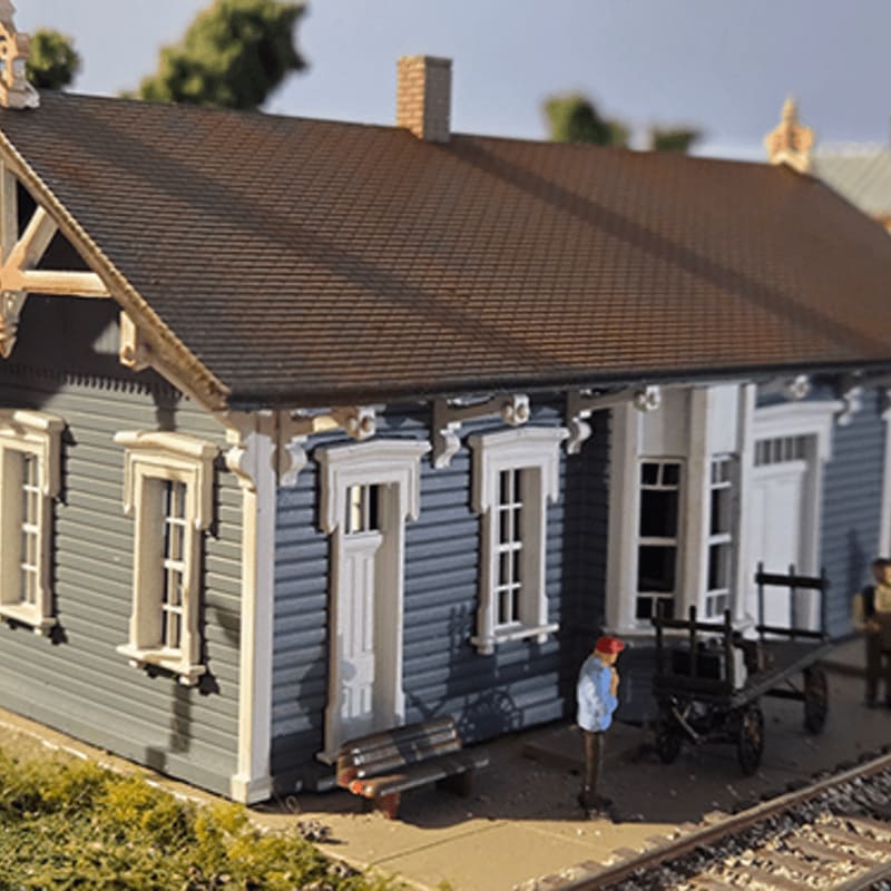 A miniature grey train station building with a brown roof stands in the foreground, set against a blurred background of green trees and a clear sky.