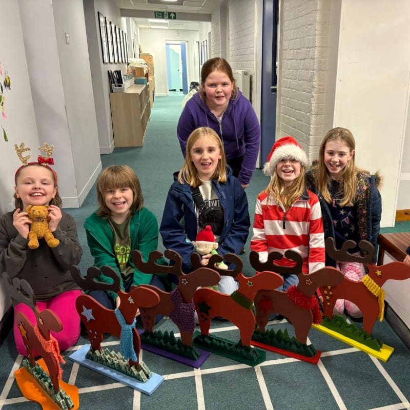 Five smiling girls sit on a hallway floor behind a row of colorful, handmade cardboard reindeer.