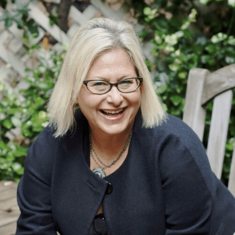 A smiling woman with blonde hair and glasses wearing a dark blazer sits in front of a lush green garden and a white lattice fence.