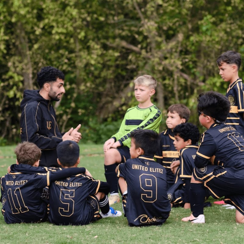 A coach in a black tracksuit talks to a group of young soccer players in black and green uniforms on a grassy field with a backdrop of lush green trees.