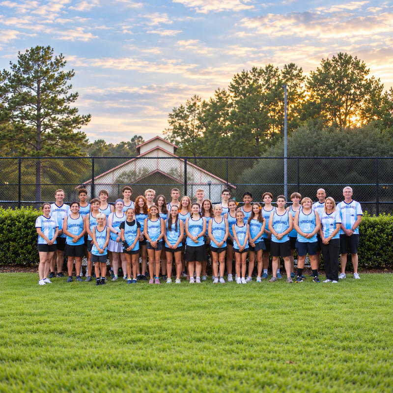 A large group of athletes in blue shirts poses on a green lawn in front of a black fence, trees, and a building under a cloudy sunset sky.
