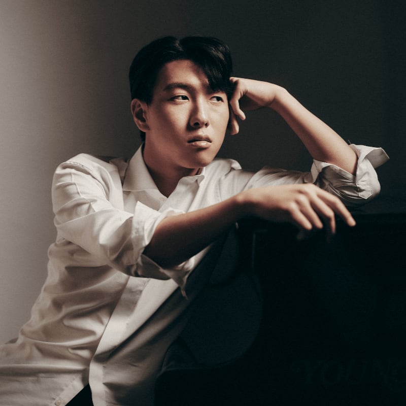 A man in a white button-down shirt leans thoughtfully against a dark surface against a muted, neutral background.